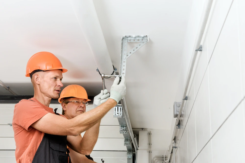 Two workers in orange hard hats tighten a bracket on a ceiling track system.